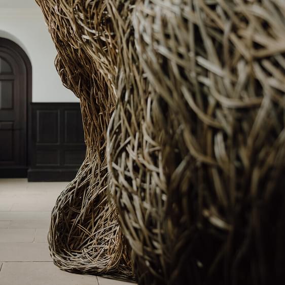 A close-up view of a large, woven sculpture made of natural materials, positioned in a spacious interior. In the background, a dark wooden door with an arched top is visible, framed by light-coloured walls and flooring.