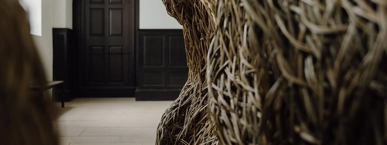 A close-up view of a large, woven sculpture made of natural materials, positioned in a spacious interior. In the background, a dark wooden door with an arched top is visible, framed by light-coloured walls and flooring.
