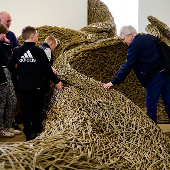 A group of four people, including two children, interact with a large, woven sculpture made of natural materials. One adult observes while another touches the textured surface. The setting appears to be a bright, spacious gallery.