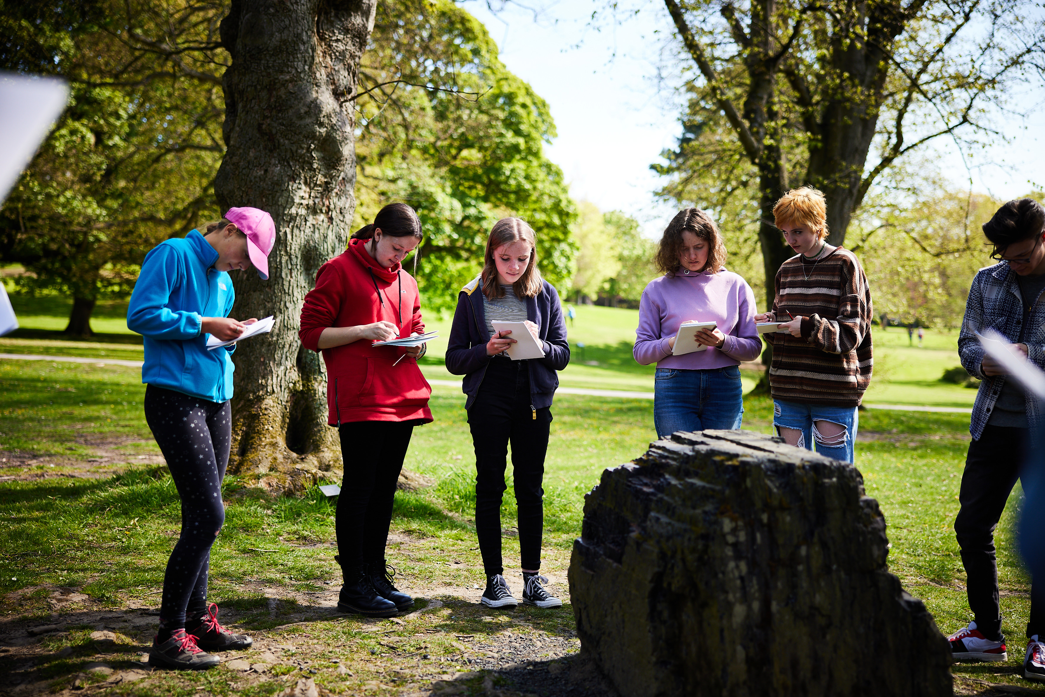 A group of young people holding sketchbooks, looking at a stone artwork in the landscape