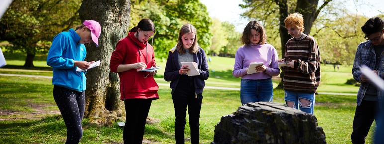 A group of young people holding sketchbooks, looking at a stone artwork in the landscape