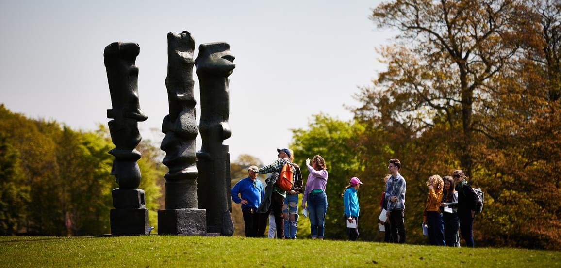 A large group of people on a tour, admiring a Henry Moore sculpture.