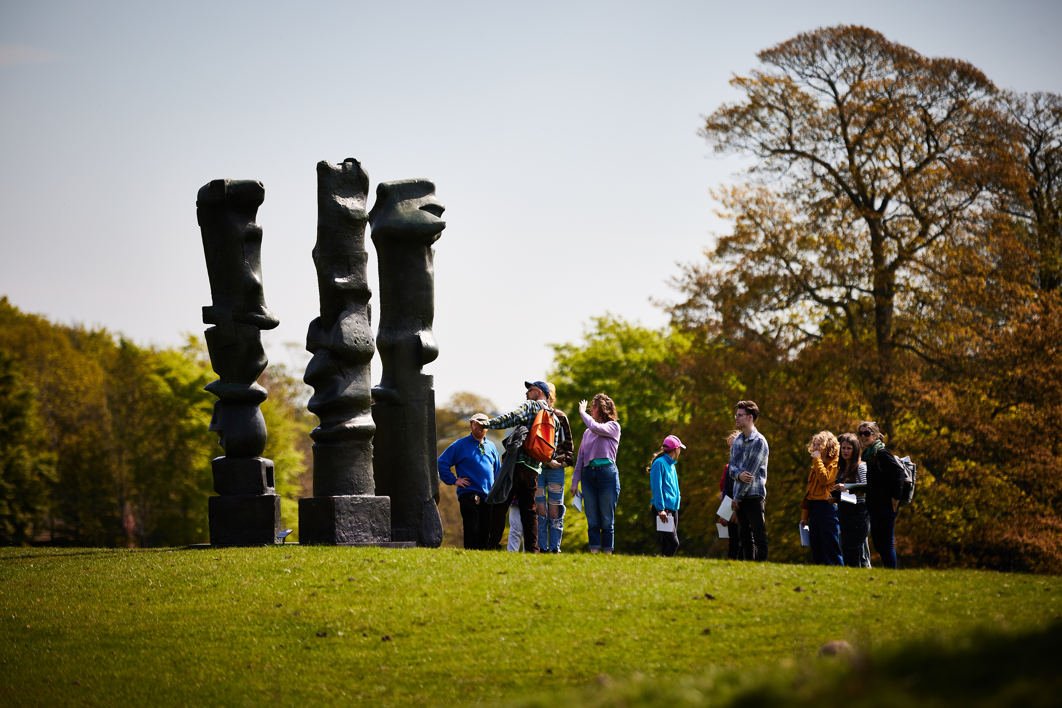 A large group of people on a tour, admiring a Henry Moore sculpture.
