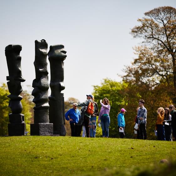 A large group of people on a tour, admiring a Henry Moore sculpture.
