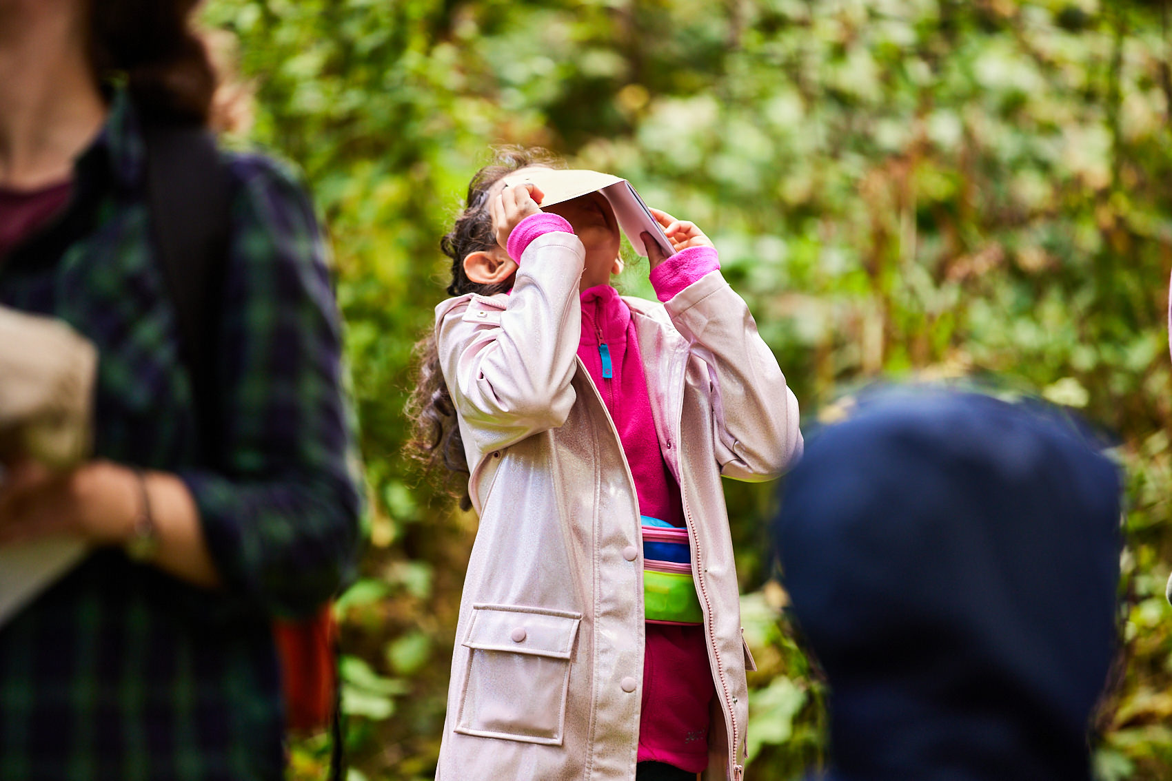 A child wearing a pink rain coat, looking through a paper viewfinder outdoors