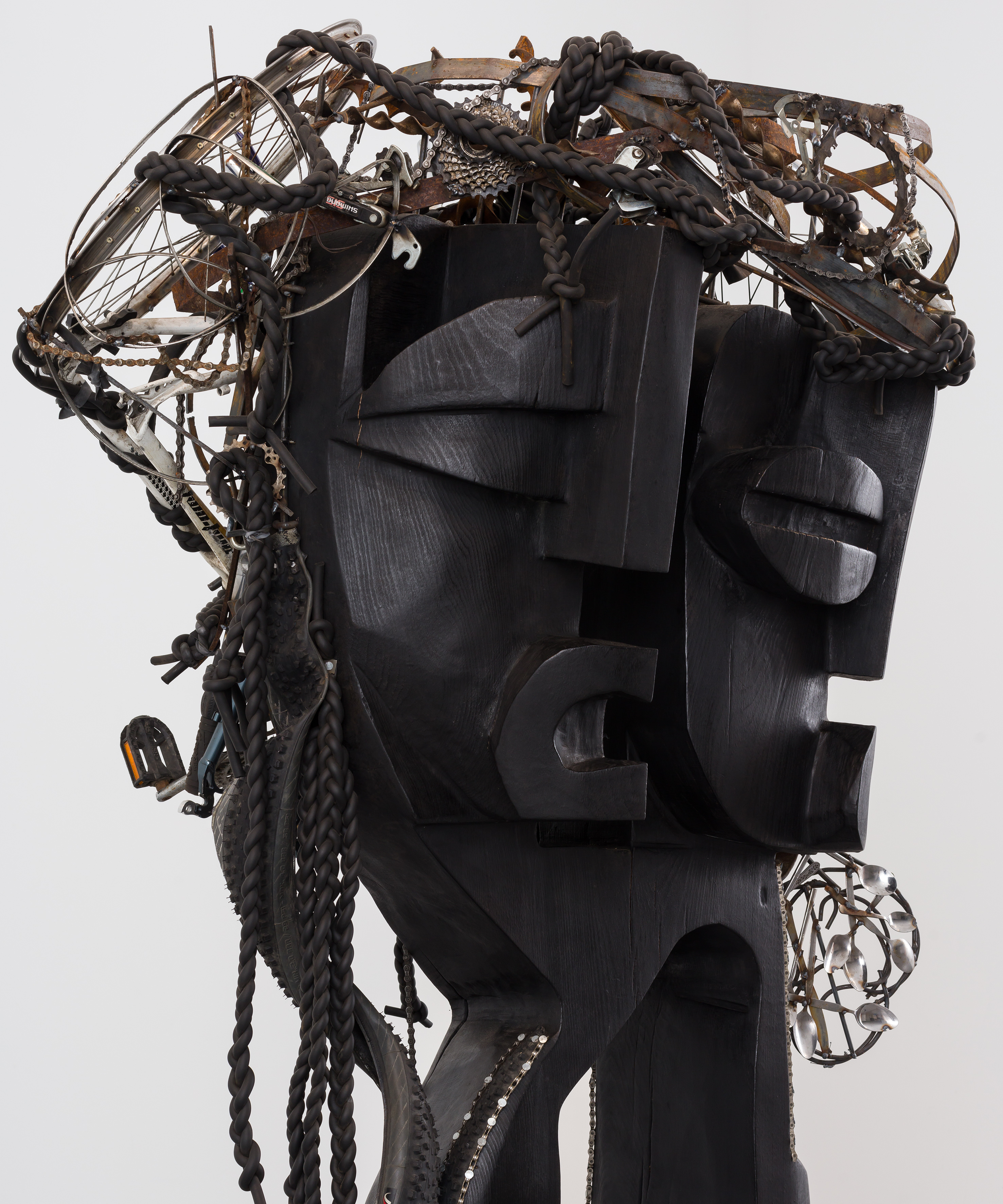 A wooden sculpture of two headed humanoid figure, with hair & accessories made from bicycle parts