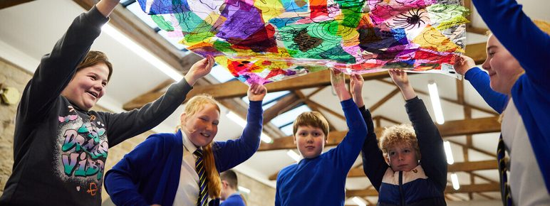 A group of children in blue school uniforms joyfully hold up a large, colourful sheet made of translucent paper. The vibrant colours and patterns create a playful atmosphere in a bright, spacious room with wooden beams.