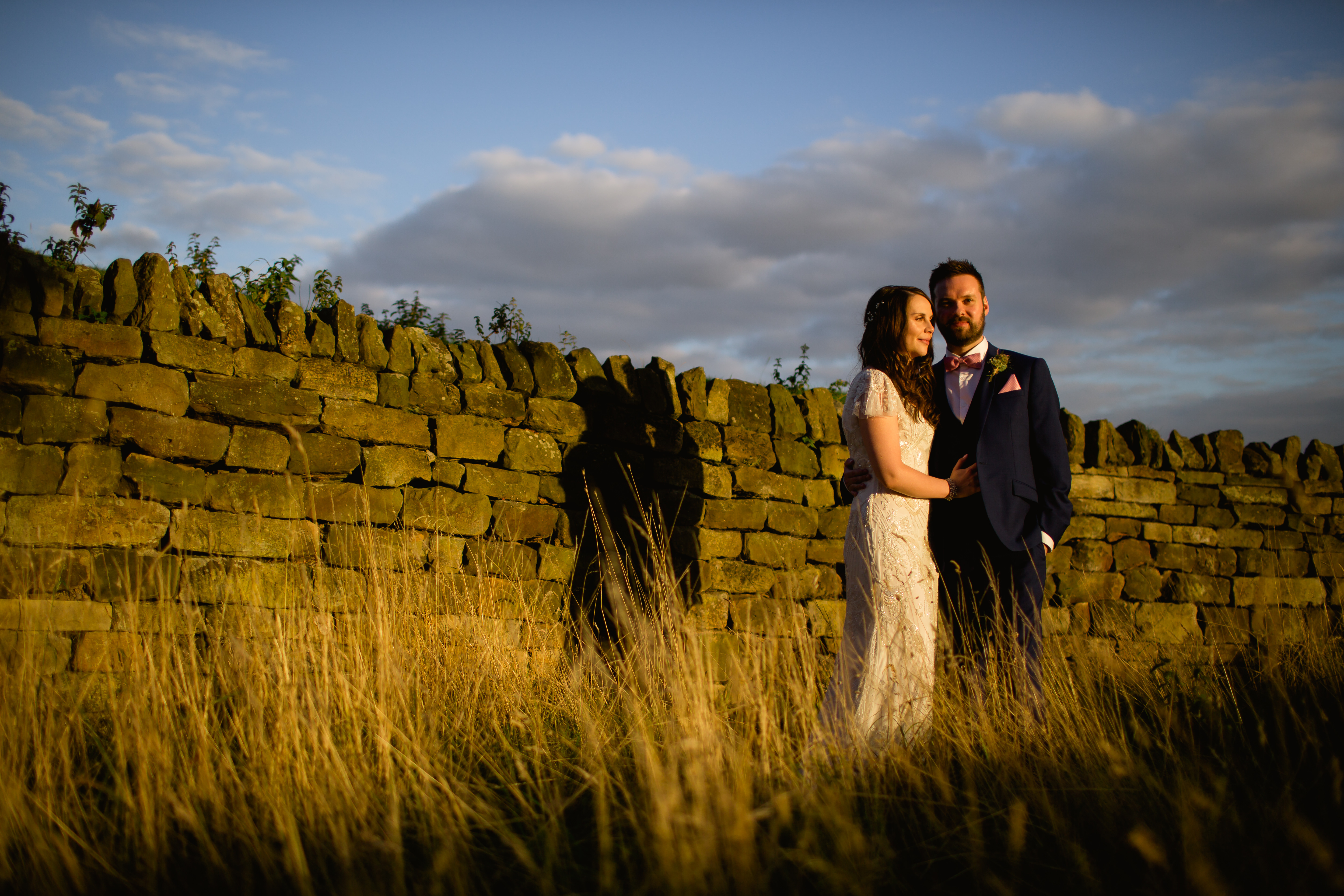 A bride and groom standing in front of a dry stone wall at sunset