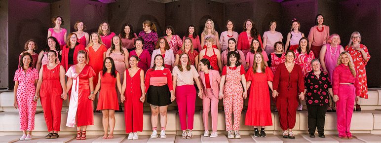 A group of women dressed in red and pink stand in rows on raised steps.