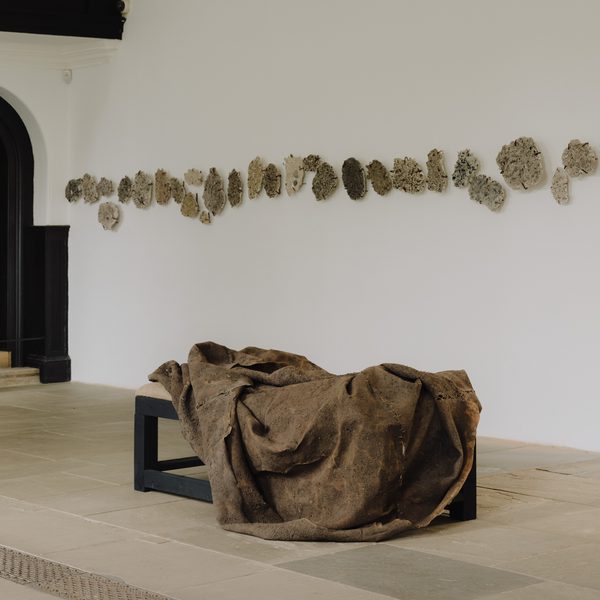 A minimalist art gallery featuring a textured, draped sculpture on a black bench. The wall behind displays a series of abstract, organic shapes in muted tones, creating a contrast with the clean, white space and dark architectural elements.