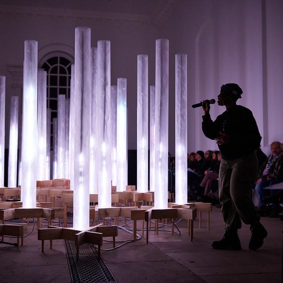 A woman singing into a microphone in front of a collection of white glowing columns