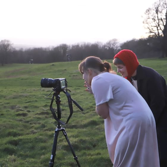 Two people, one wearing a red woolly hat, the other a nightdress crouched over looking into the lense of a camera.