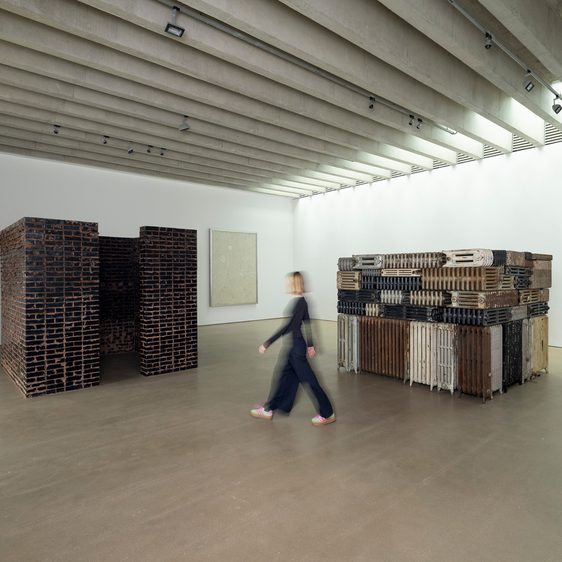 Two large sculptures in a gallery and a work affixed to the wall, with a woman walking in between.