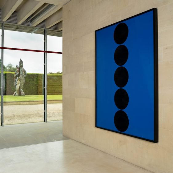 A blue work with black dots hung on a gallery wall, in the outside through the window is a figurative sculpture.