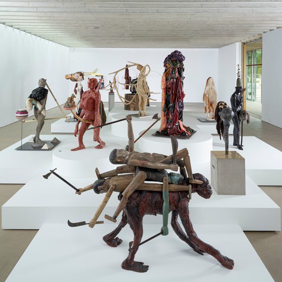 A display of human/animal figures on white plinths inside a gallery space.