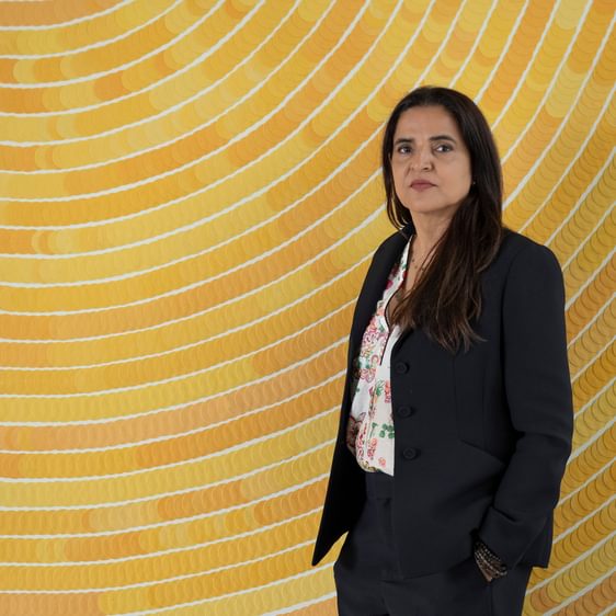 A woman wearing a black suit standing in front of a yellow spiral wall-based artwork.