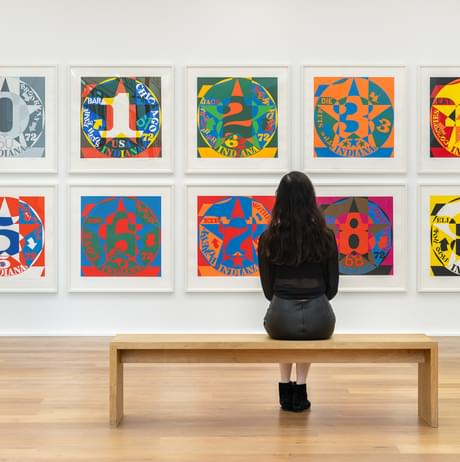 A woman sitting on a bench in front of a display of brightly coloured pop art prints in a gallery.