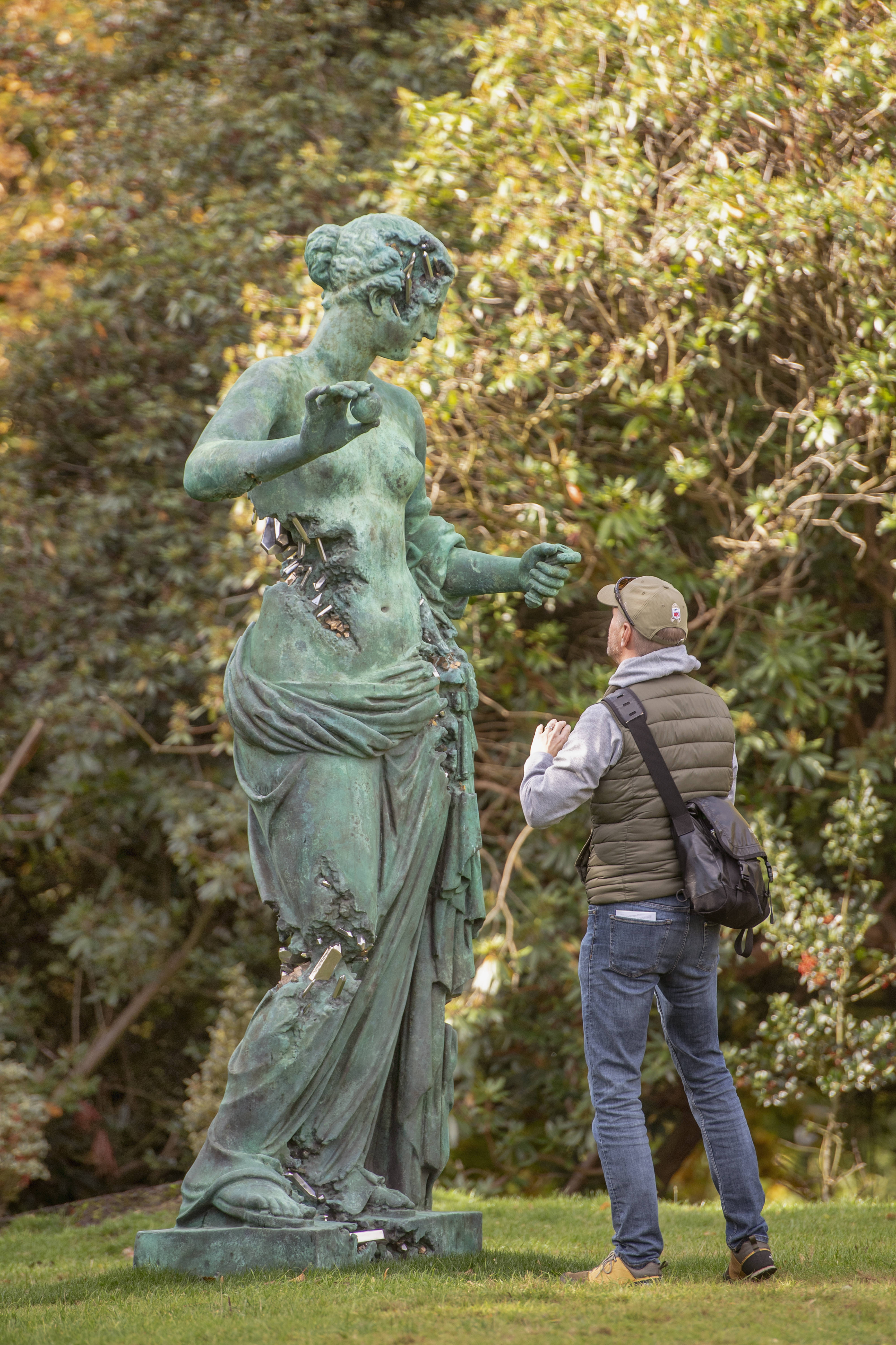 A man taking a photograph of a giant bronze sculpture of a naked woman, with eroded bronze crystallised sections.