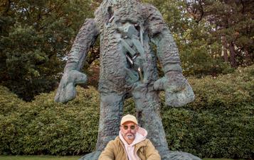 The artist Daniel Arsham, wearing a winter coat and sunglasses, sitting on the base of a giant rabbit sculpture.