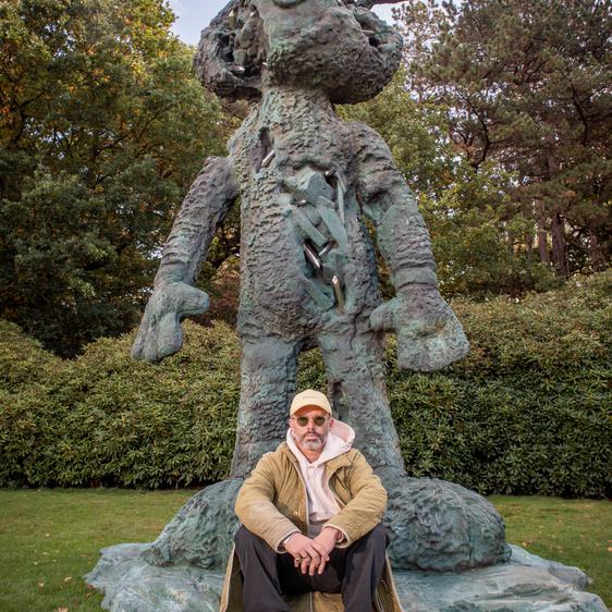 The artist Daniel Arsham, wearing a winter coat and sunglasses, sitting on the base of a giant rabbit sculpture.