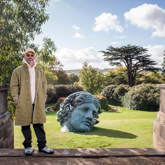 A man wearing a tan jacket and baseball cap, standing in front of a sculpture of a woman's head with eroded crystal sections.