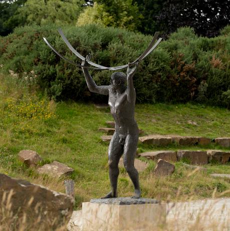 A bronze sculpture of a naked male figure holding curved bars above his head, displayed outdoors.