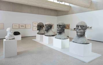 Four large head sculptures displayed on plinths in a row inside a gallery space.