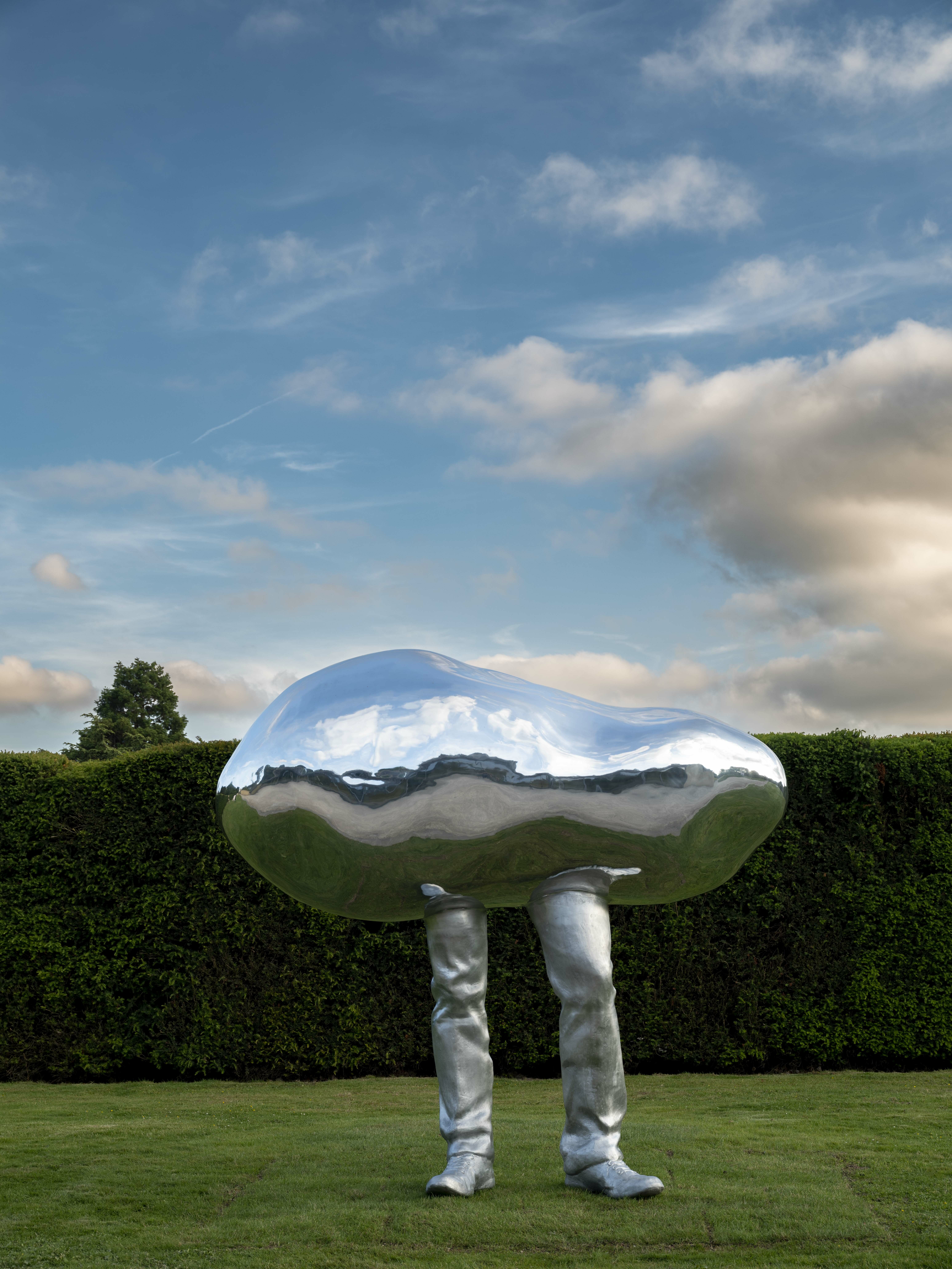 A silver pebble with legs sculpture, stood on grass underneath a blue sky with grey clouds