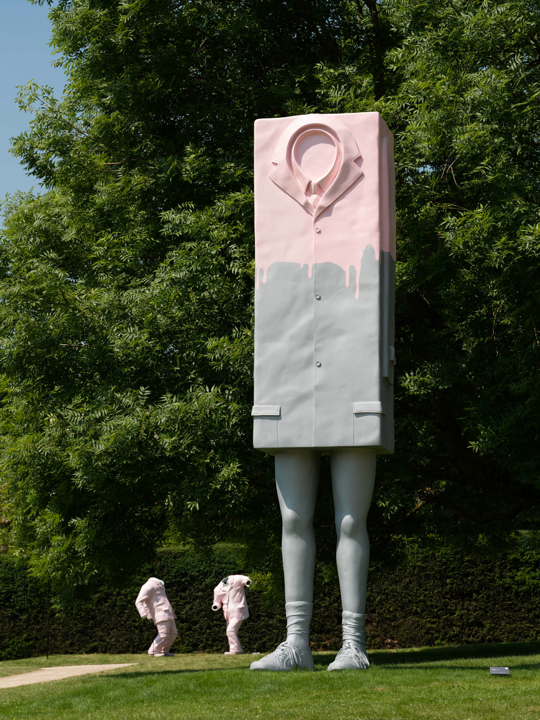 A pink and grey box on legs wearing a suit, two headless sculptures wearing pink suits in the background