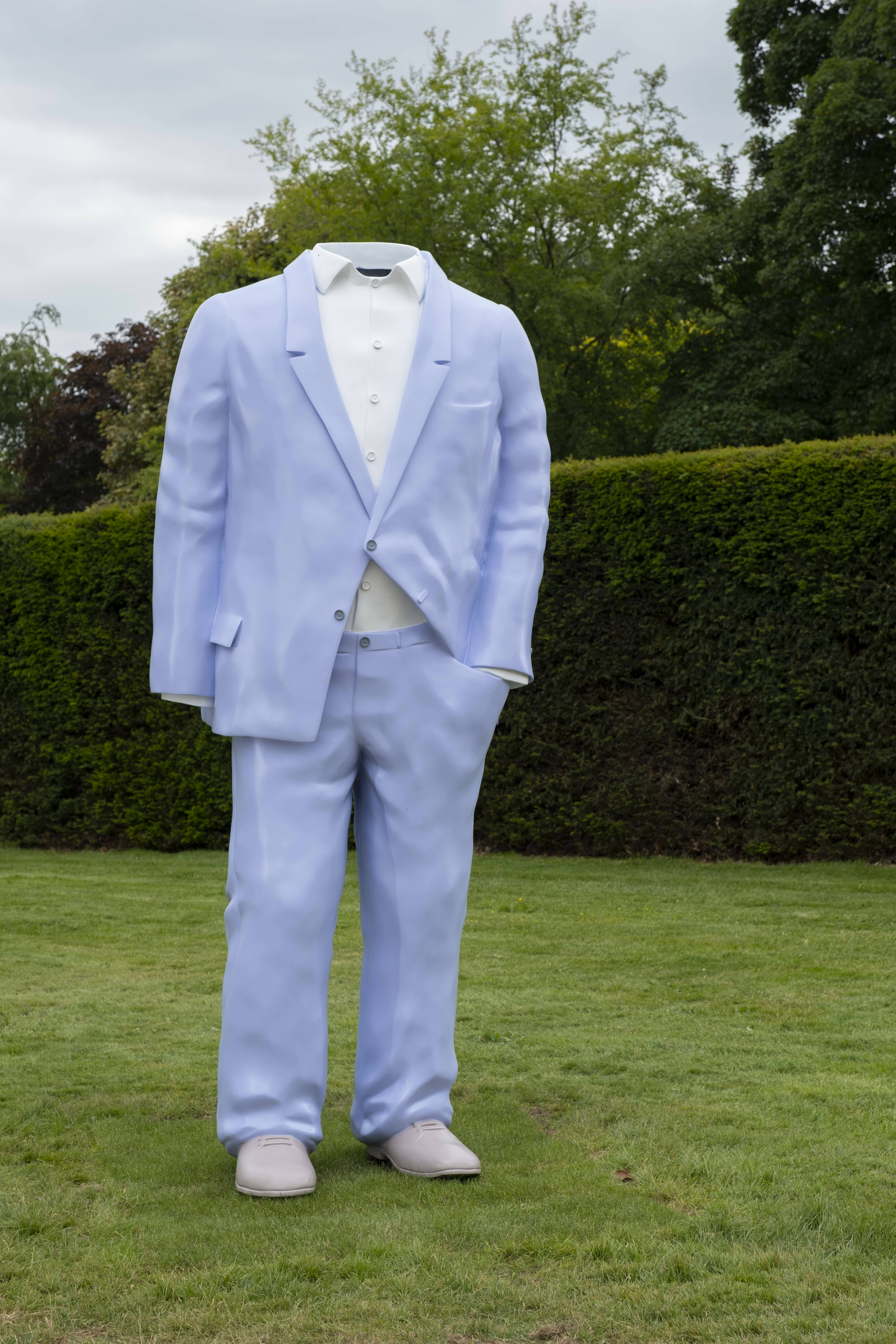 Sculpture of a headless person dressed in a purple suit and white shirt stood on grass.