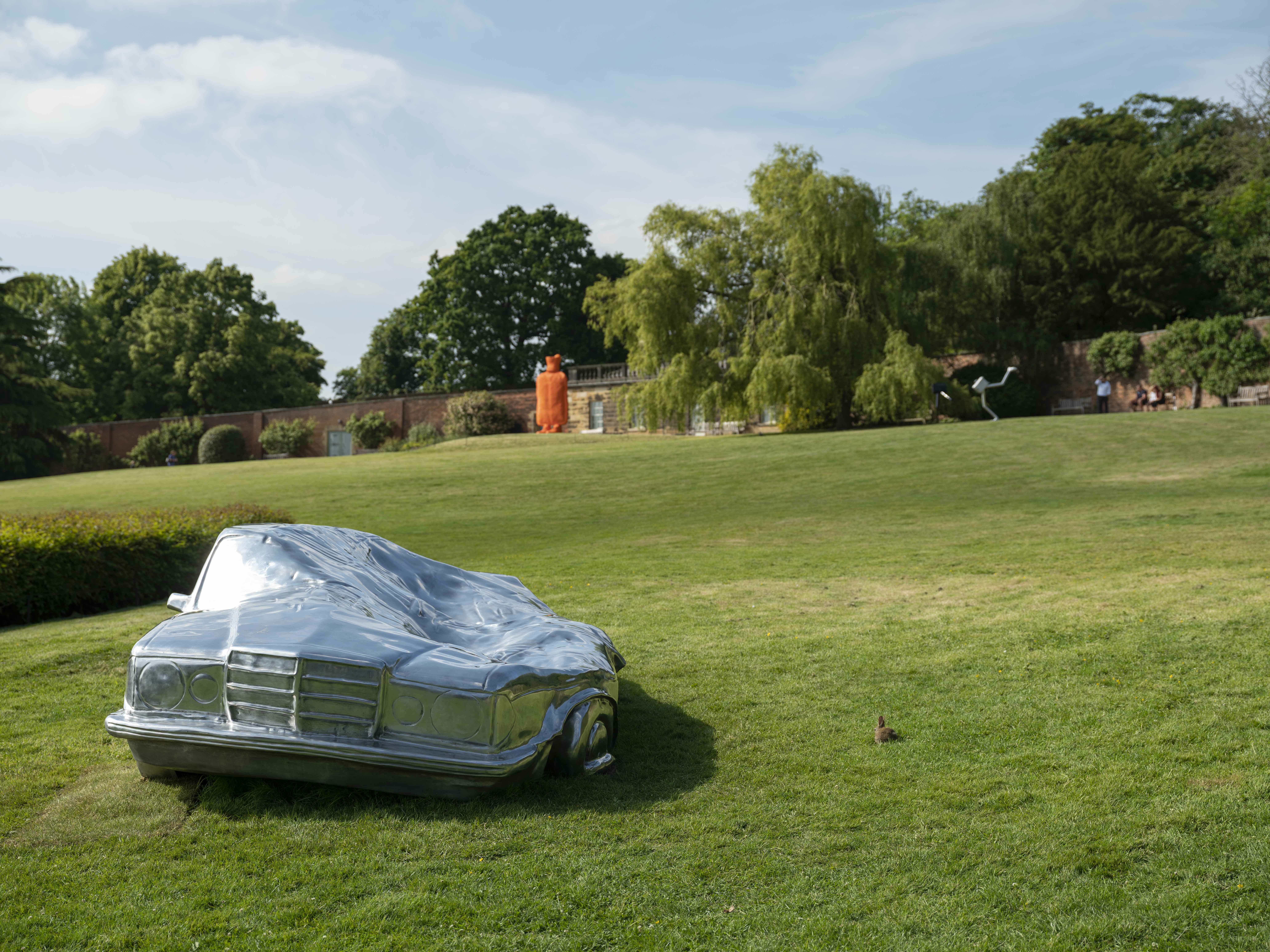 A silver squashed car on the lawn and a orange hot water bottle in the distance