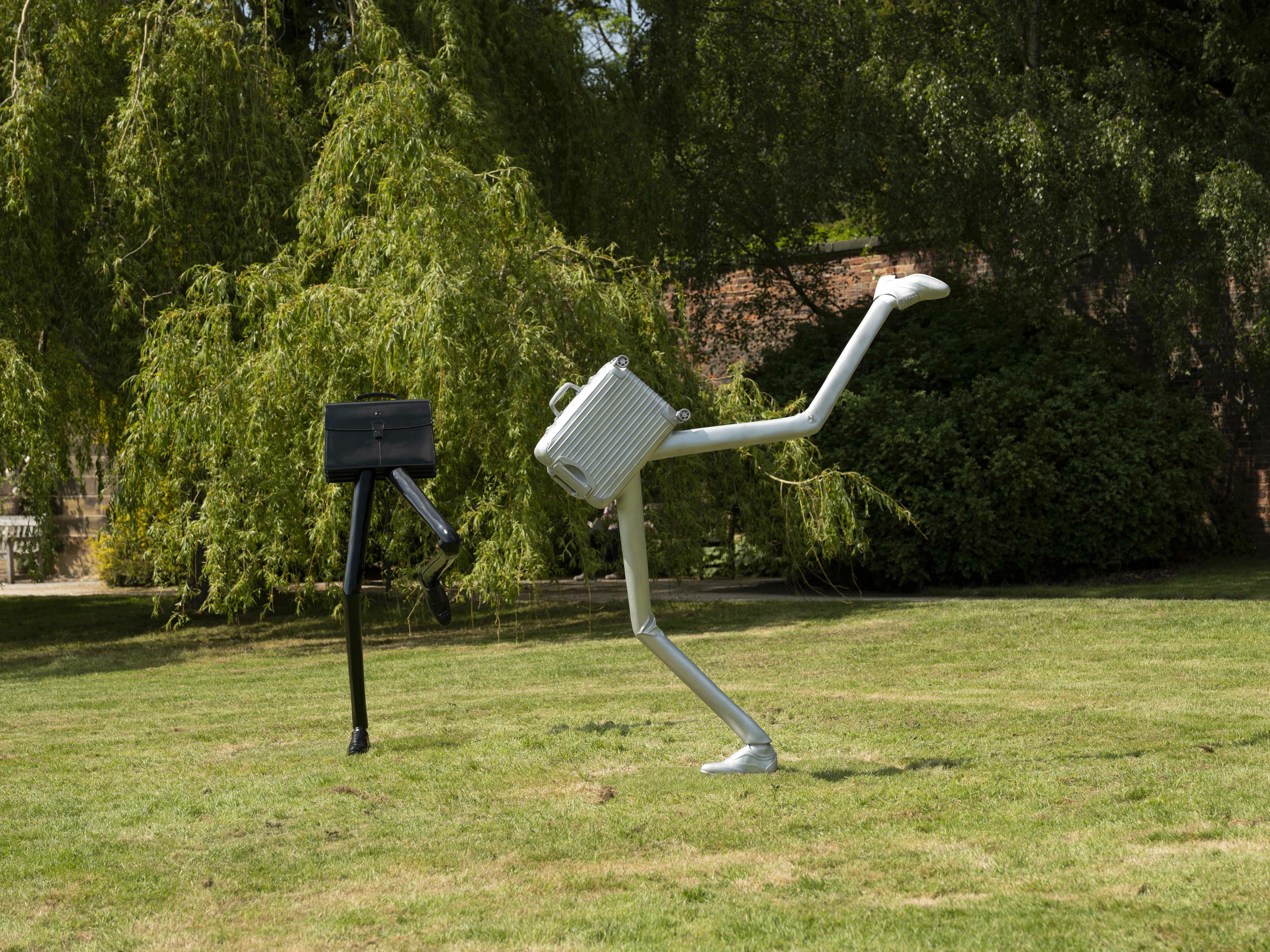 A silver suitcase sculpture and black briefcase sculpture both with legs on a lawn.