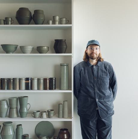 Man with shoulder length hair, glasses and a cap, leaning on shelves stacked with ceramic vessels.