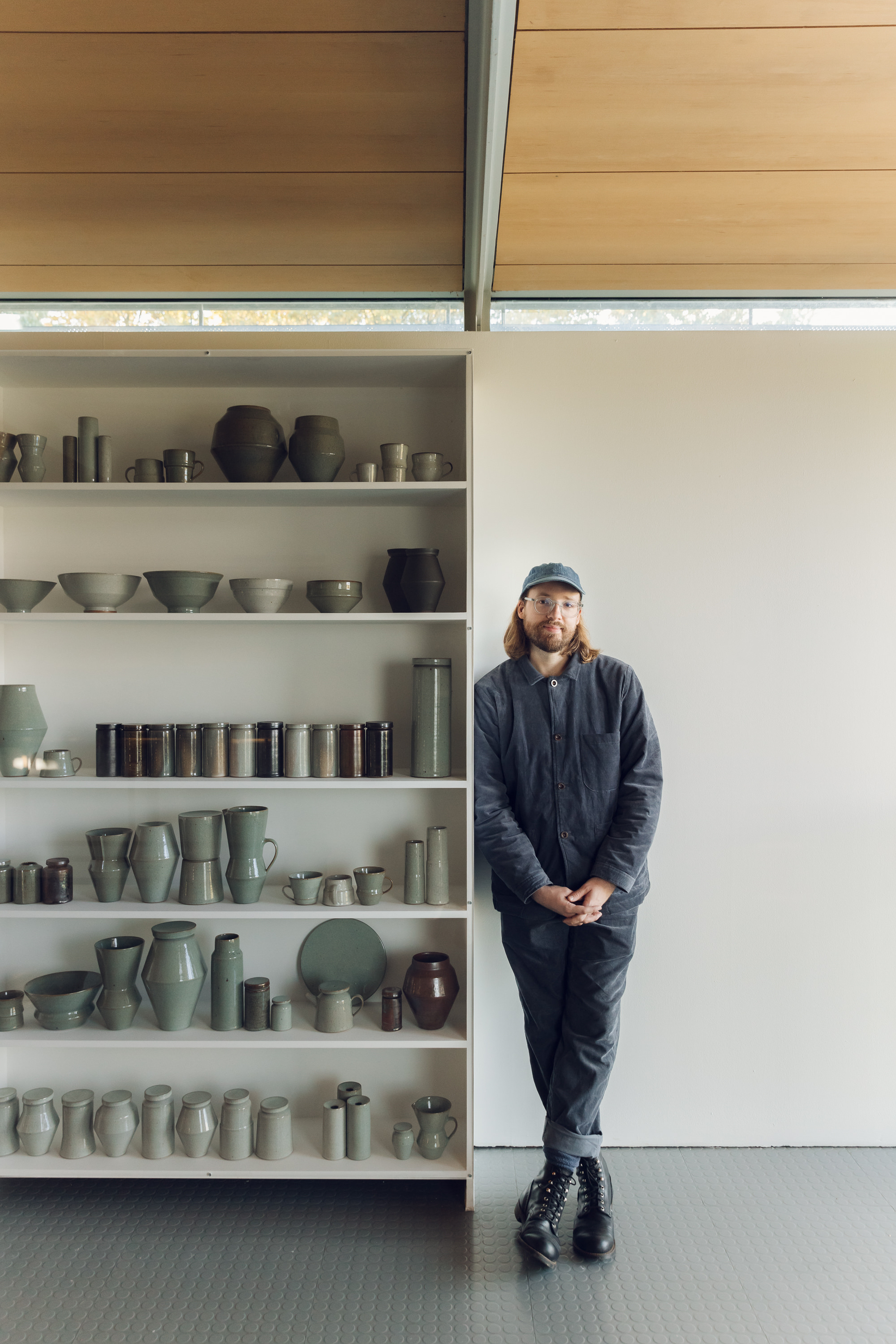 Man with shoulder length hair, glasses and a cap, leaning on shelves stacked with ceramic vessels.