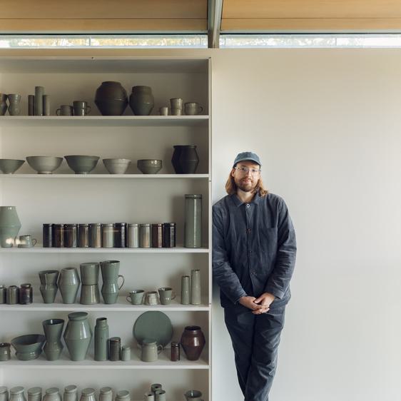 Man with shoulder length hair, glasses and a cap, leaning on shelves stacked with ceramic vessels.