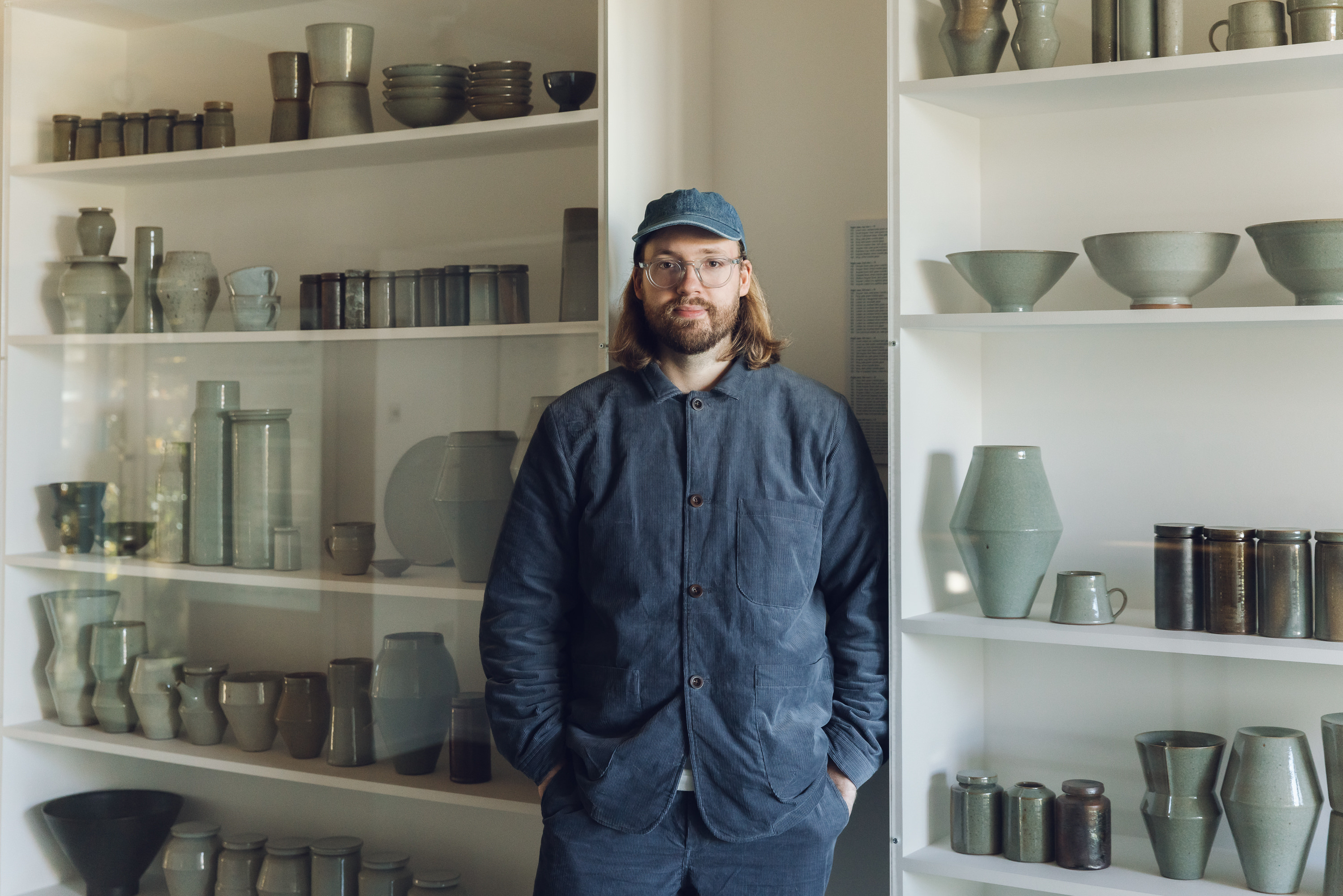 Man with shoulder length hair wearing glasses and a cap standing in front of shelves packed with ceramic vessels