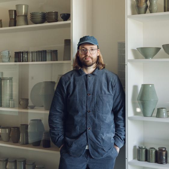 Man with shoulder length hair wearing glasses and a cap standing in front of shelves packed with ceramic vessels