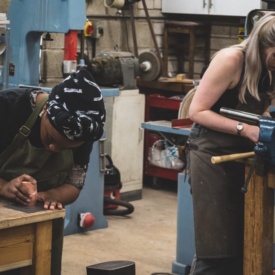 Two women working in a workshop, one at a wooden table and one on a machine.
