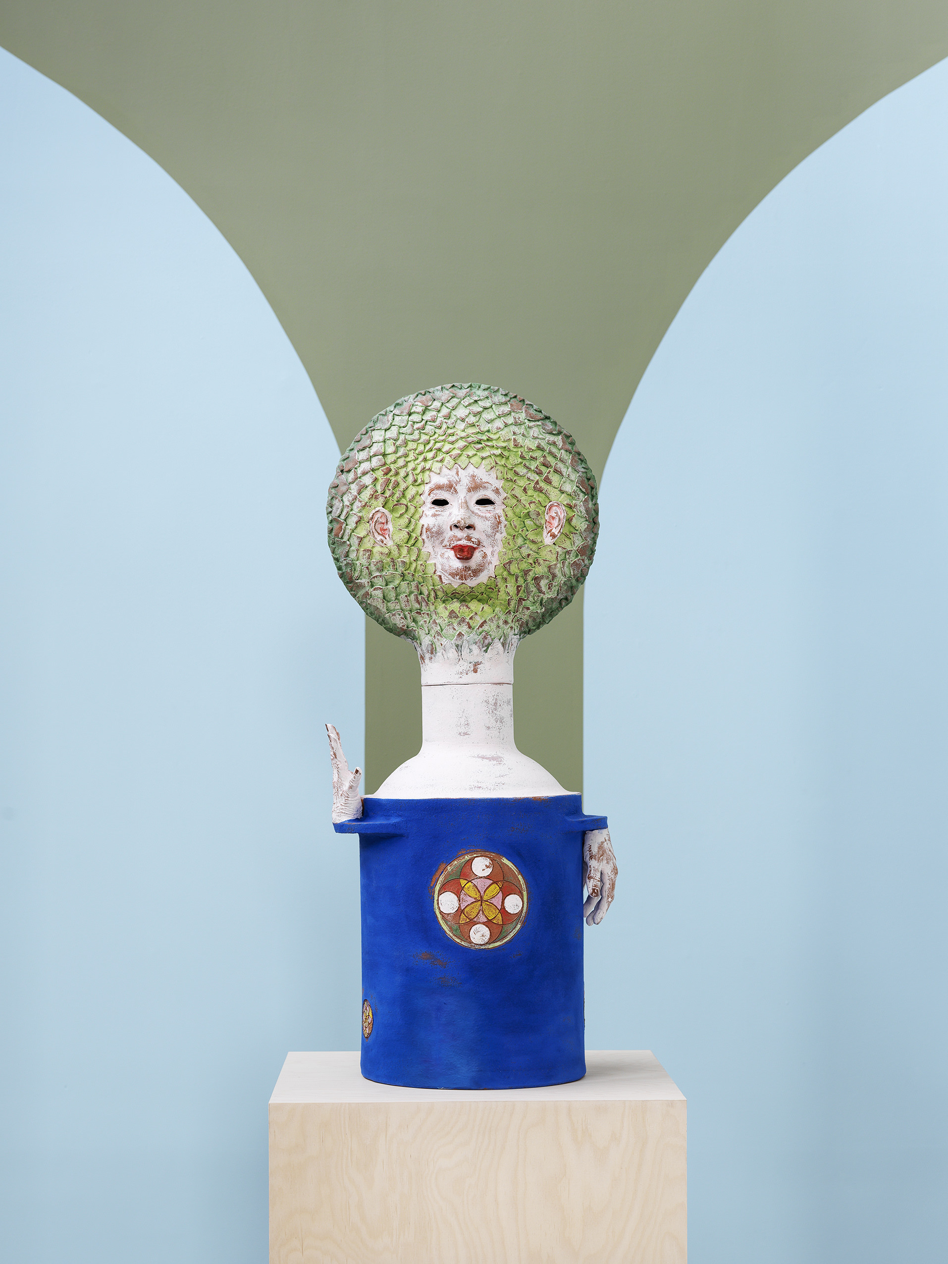 Sculpture on a plinth imitating a flower, with a blue pot and a green face, sticking its tongue out