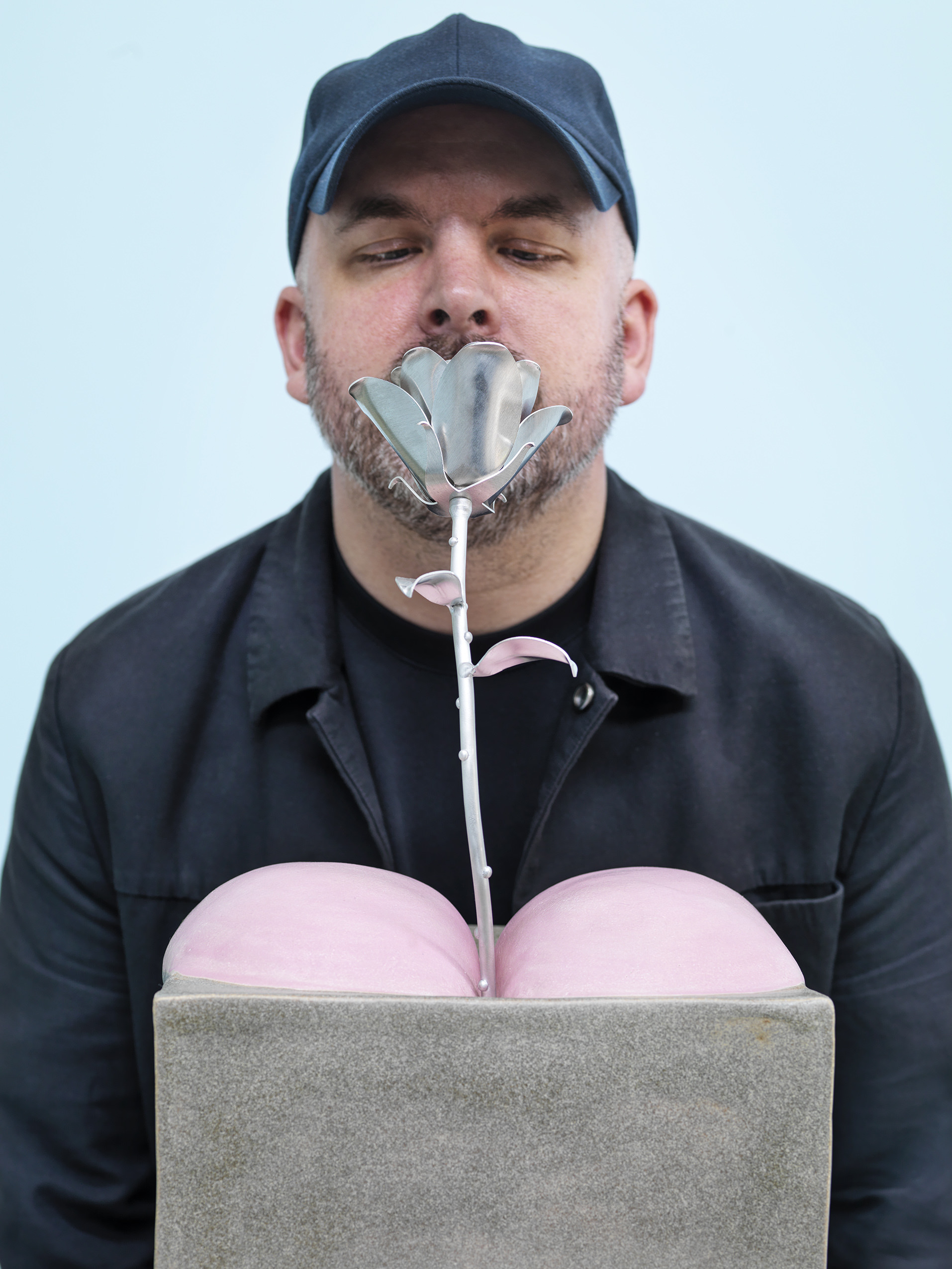 Man smelling a silver rose growing between sculpture of two pink bottom cheeks