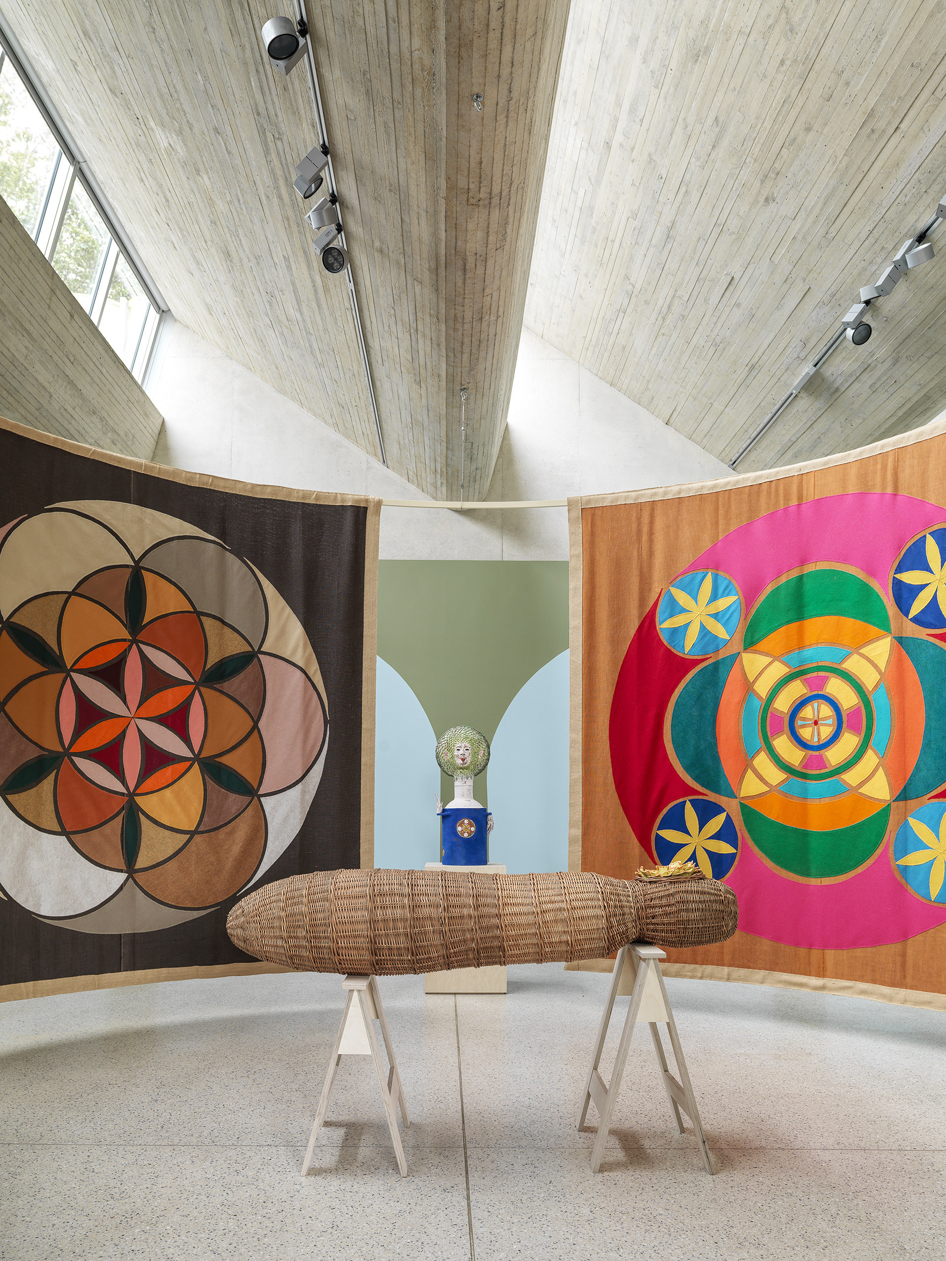 Indoor gallery space with two hanging tapestries and a woven basket coffin