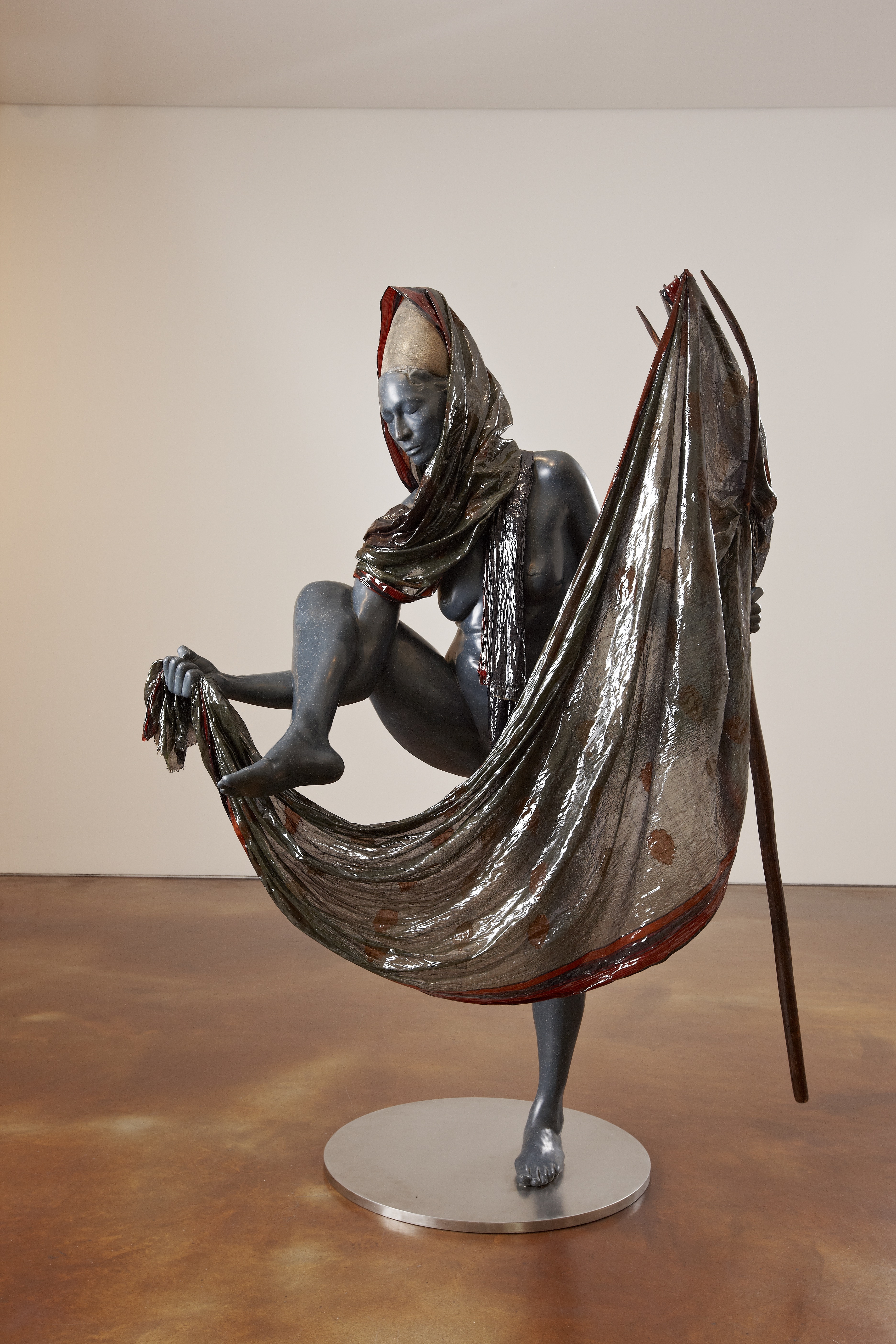 Sculpture of a woman poised on one leg, the other in the air, draped in a silver sari.