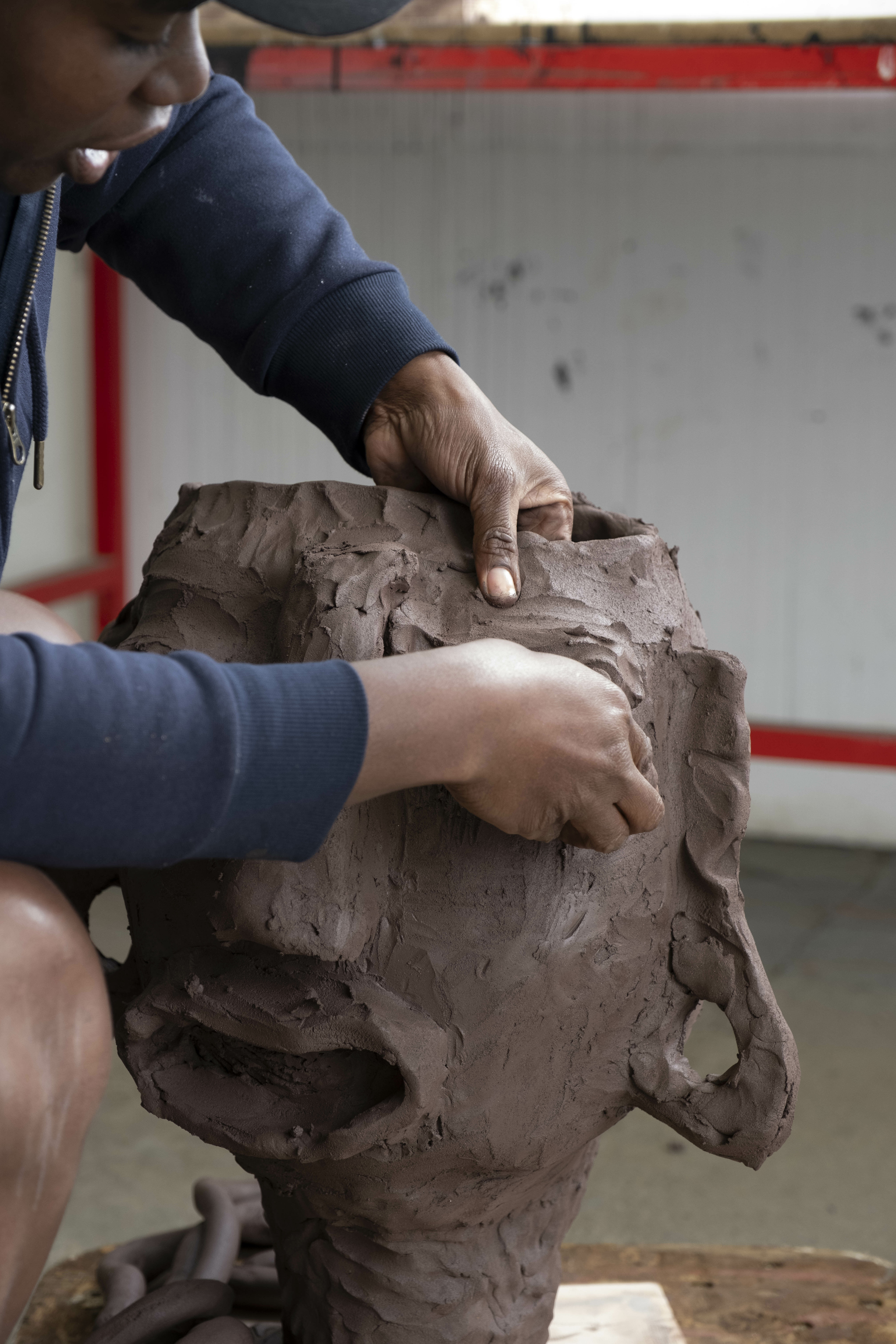 A black woman wearing shorts and a sweatshirt, working on a clay head sculpture