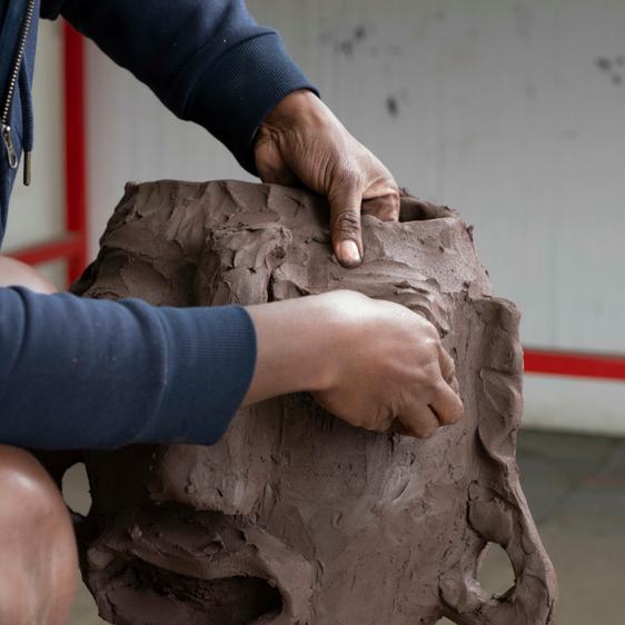 A black woman wearing shorts and a sweatshirt, working on a clay head sculpture