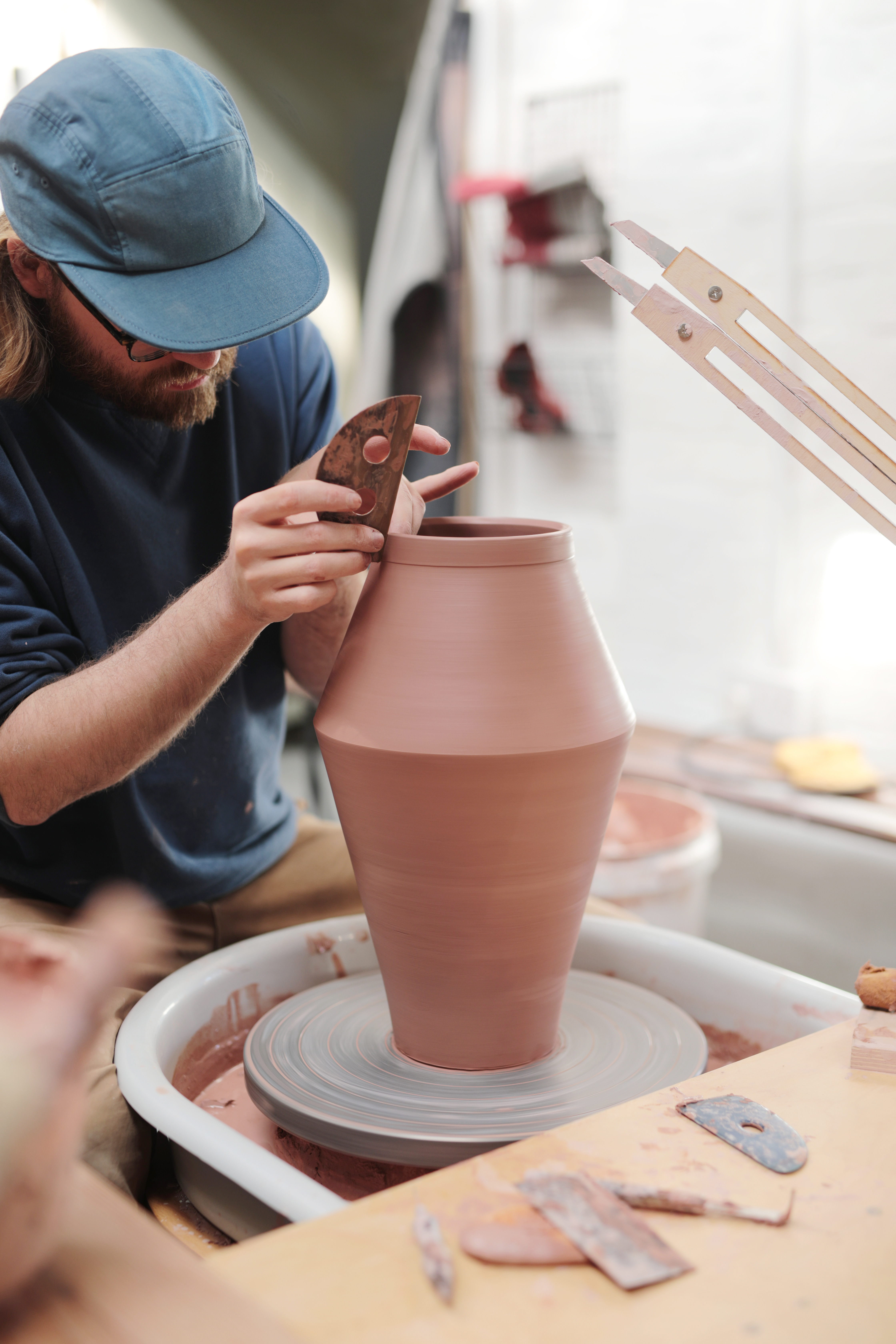 A man wearing a blue cap, trimming the rim of a large pot on a potter's wheel.