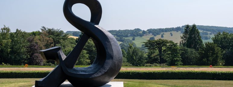 A large, abstract black sculpture resembling an ampersand stands on a grassy area, with rolling green hills and trees in the background under a clear blue sky. The sculpture's smooth curves contrast with the natural landscape.