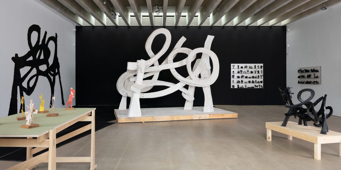 An art gallery featuring a large white abstract sculpture with looping forms at its centre. Surrounding it are smaller sculptures in various colours and styles displayed on wooden tables, against a contrasting black wall.