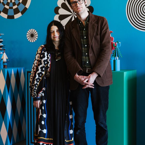 A man in glasses and a brown jacket stands beside a woman in a patterned dress, abstract shapes on a blue background behind them. The scene is set in a gallery, showcasing contemporary art.