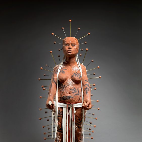 A life-sized sculpture of a figure covered in intricate body art and clay, standing confidently. The figure is adorned with numerous pins radiating from the head, and white ribbons hang from the waist, set against a dark background.
