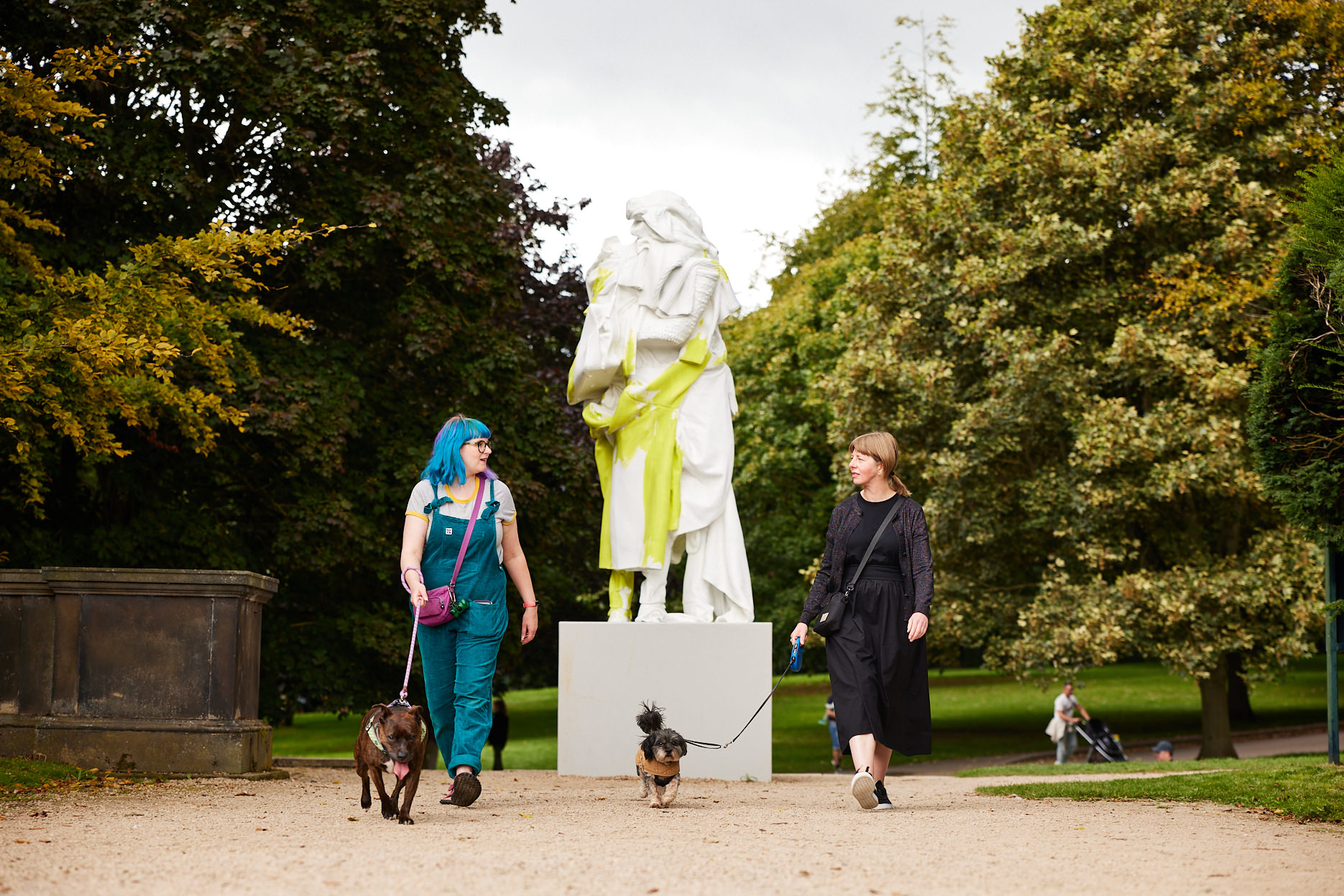 Two women walking dogs on leads past a tall white human-shaped sculpture